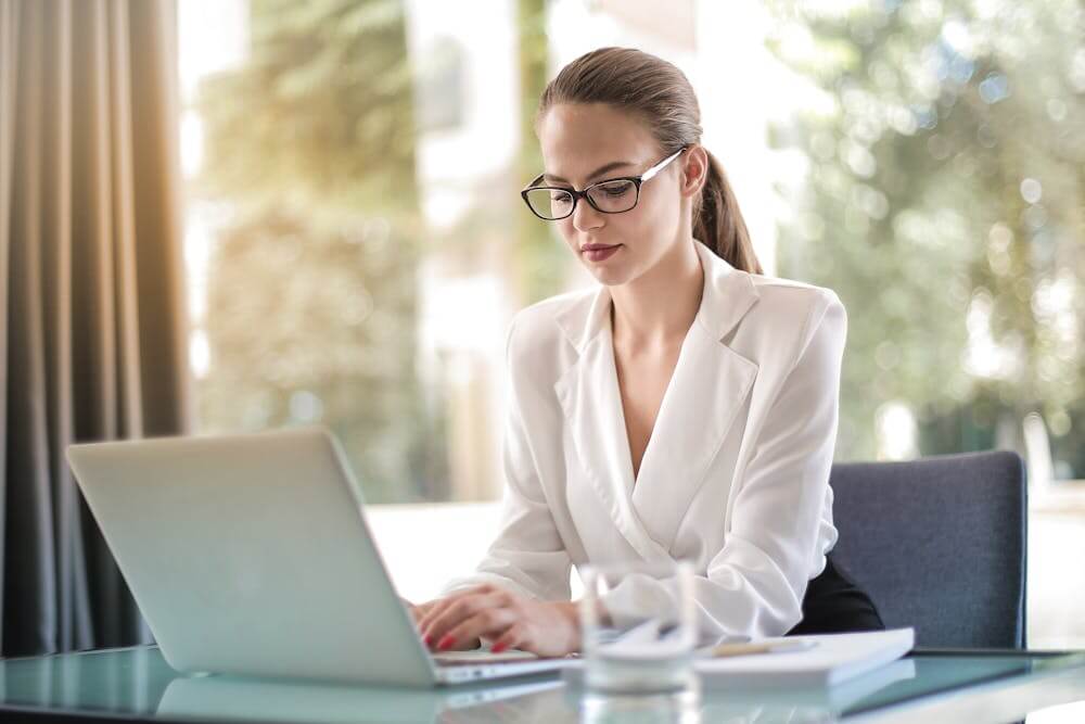 A businesswoman in glasses types on a laptop at a desk.